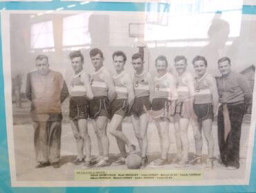 photo en noir et blanc sur laquelle posent 6 joueurs de basket en tenue de sport, entourés de deux personnes en tenue de ville