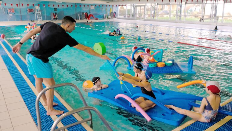 Un étudiant en Staps tend un objet en mousse à une enfant pour qu'elle construise un radeau qui flotte dans la piscine municipale.