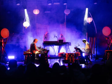 Vue de la scène d'un concert : 4 musiciens masqués, debout devant des consoles électroniques. on aperçoit le public de dos.